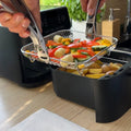 Stackable stainless steel air fryer basket lifted with tongs from black air fryer drawer filled with roasted vegetables