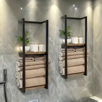 Two black metal wall towel racks with wooden shelves mounted on grey marble bathroom tiles, rolled beige towels and small green plant styled on upper shelf