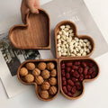 Four heart-shaped solid walnut wooden snack bowls arranged in clover-leaf formation filled with nuts and dried fruit on white bench