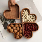 Four heart-shaped solid walnut wooden snack bowls arranged in clover-leaf formation filled with nuts and dried fruit on white bench