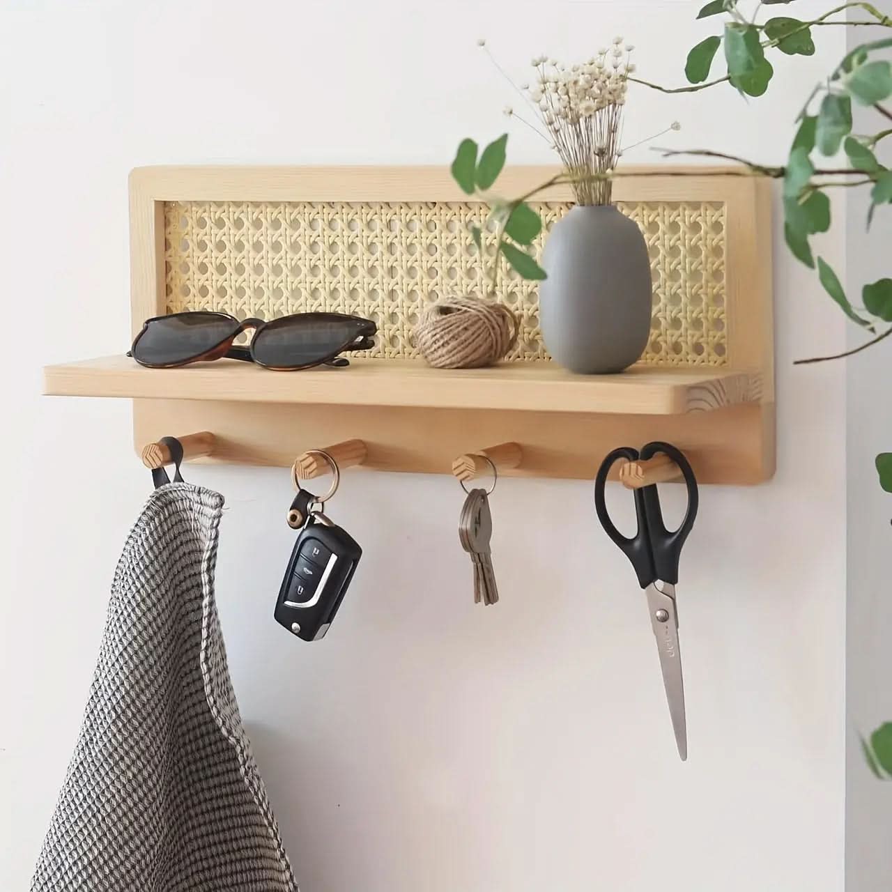 Wooden wall shelf with rattan back panel and four hooks mounted in hallway, keys and cloth hanging, vase on top ledge