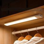 Motion sensor LED light mounted under wooden wardrobe shelf illuminating hanging clothes