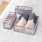 Three grey nylon mesh drawer organisers holding bras, underwear, and socks in separate compartments on white bed linen