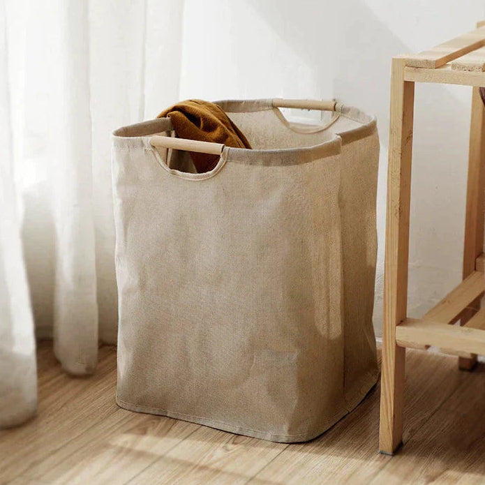 Foldable beige cotton-linen laundry basket with wooden handles on timber floor in bright bedroom lifestyle setting