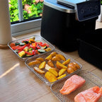 Three stainless steel mesh air fryer baskets in square, rectangular, and round shapes on wooden bench next to black air fryer