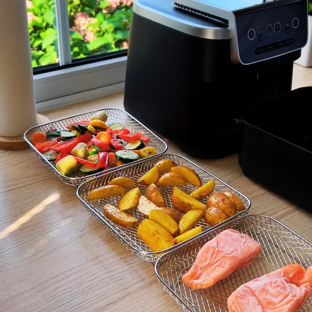 Three stainless steel mesh air fryer baskets in square, rectangular, and round shapes on wooden bench next to black air fryer
