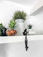 Abstract figure sculpture in black resin sitting on a white shelf styled with small indoor potted plants and a ceramic white vase
