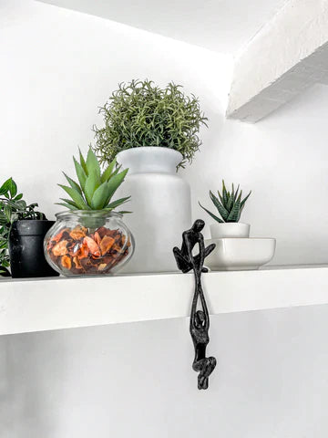 Abstract figure sculpture in black resin sitting on a white shelf styled with small indoor potted plants and a ceramic white vase
