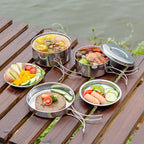 Stainless steel camping pots and pans with folding wire handles arranged on a timber slatted table beside a river