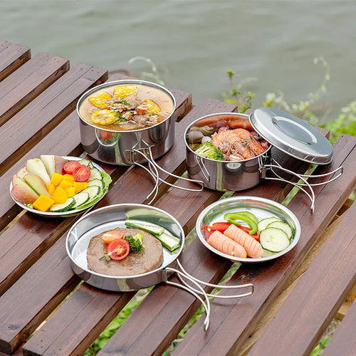 Stainless steel camping pots and pans with folding wire handles arranged on a timber slatted table beside a river