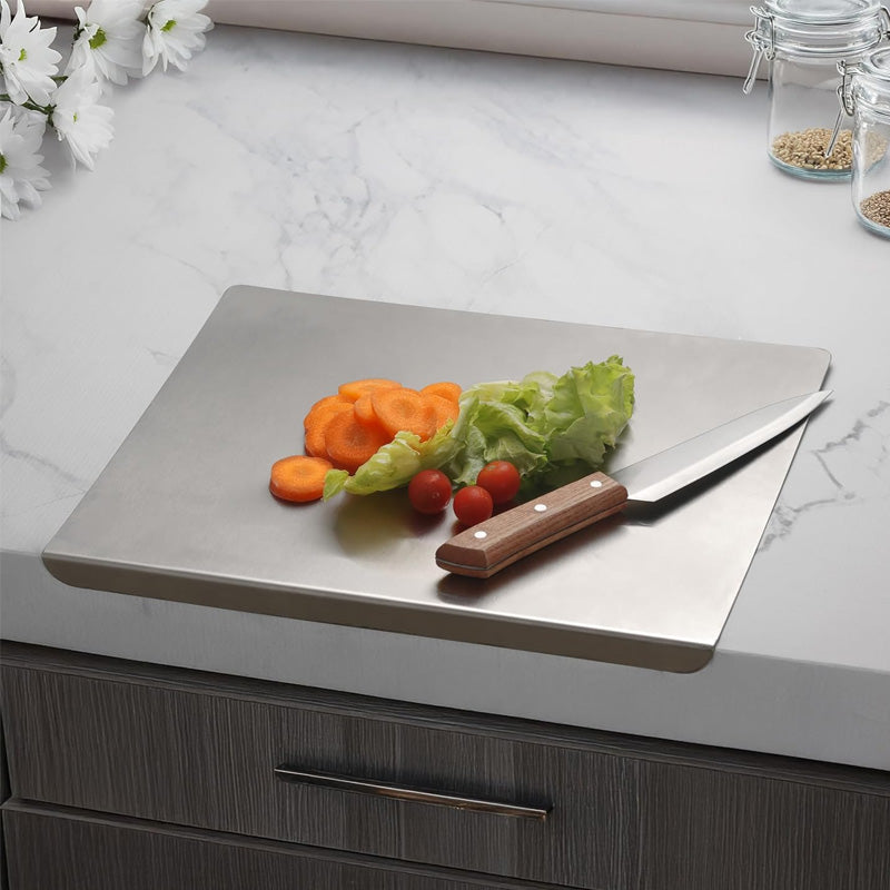 Hands cutting vegetables on stainless steel board with edge hooked over kitchen benchtop