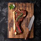 Teak cutting board with grilled steak garnished with rosemary and chilli peppers beside kitchen knife