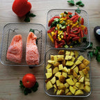 Stainless steel air fryer baskets loaded with potato wedges, salmon, and vegetables displayed on kitchen bench in natural light