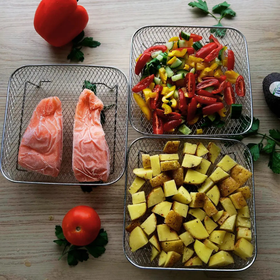 Stainless steel air fryer baskets loaded with potato wedges, salmon, and vegetables displayed on kitchen bench in natural light